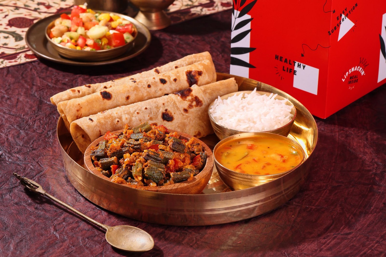 Milli Meals thali with rotis, bhindi masala, dal, rice and salad on a brass plate beside a red Milli Meals box